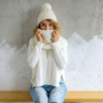 beautiful young woman sitting against wall wearing white sweater knitted hat scarf